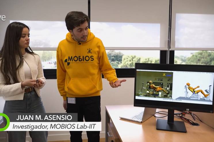 A man in a yellow hoodie and a woman stand discussing over a computer monitor displaying graphical animations.