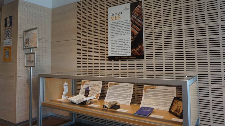 La imagen muestra una exposición con varios libros y objetos en una vitrina, junto a un panel informativo sobre la 'Pieza del Mes'.