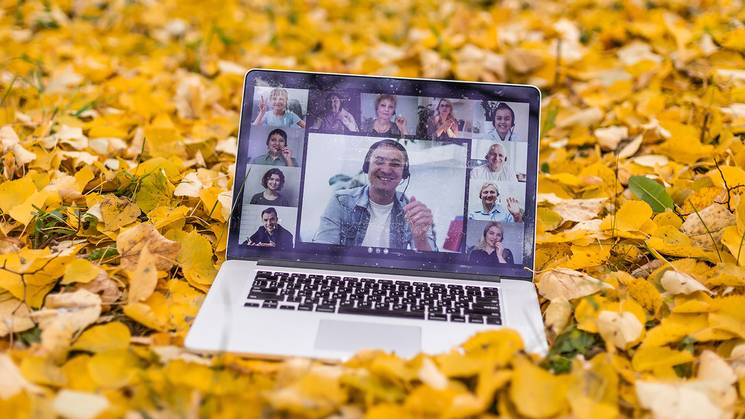 Una computadora portátil abierta sobre un lecho de hojas amarillas muestra una videoconferencia con varias personas sonrientes.