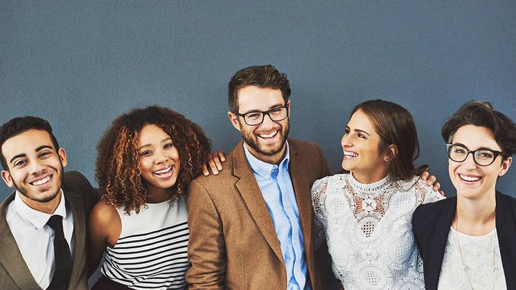 Un grupo de cinco jóvenes sonrientes posando juntos frente a una pared gris.