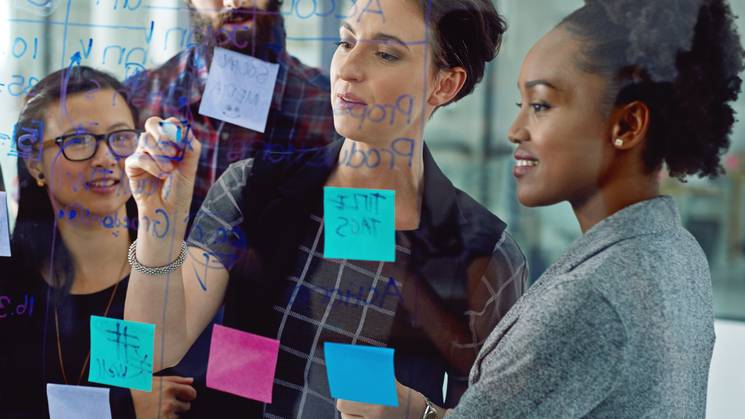 A group of four diverse colleagues brainstorming with sticky notes on a glass wall in a modern office.