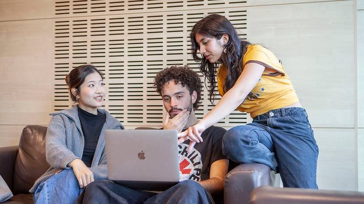 Tres personas están sentadas en un sofá, mirando una computadora portátil y conversando entre ellas.