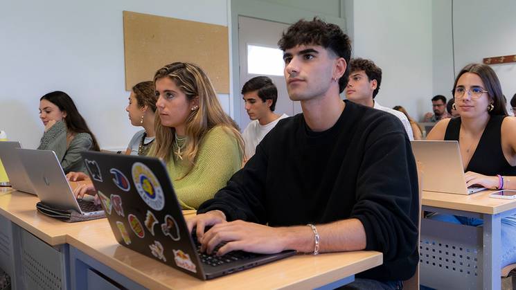 Un grupo de estudiantes está sentado en un aula con laptops, prestando atención a una clase.