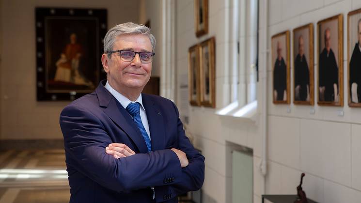 A man in a blue suit and glasses stands with his arms crossed in a corridor lined with portrait paintings.