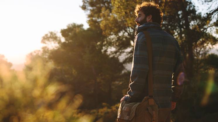 Un hombre con barba se encuentra en un bosque al atardecer, mirando hacia el horizonte.