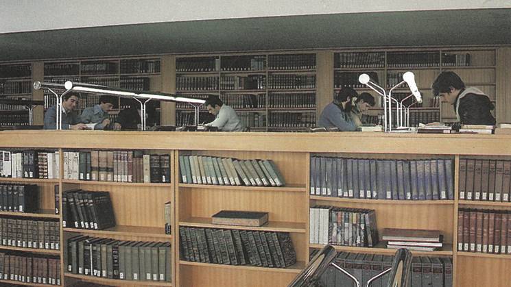 People studying in a library with desks, lamps, and bookshelves filled with books.