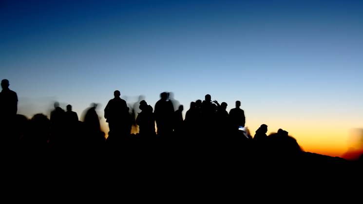 Silhouettes of people gathered outdoors at sunset with a vibrant sky in the background.