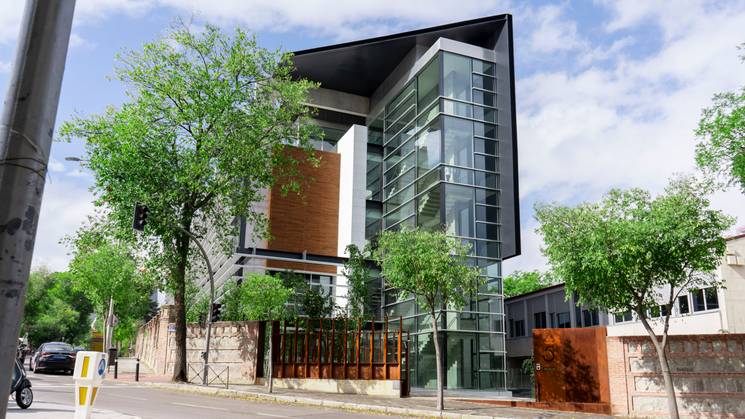 Modern glass building with orange accents on a city street lined with green trees.