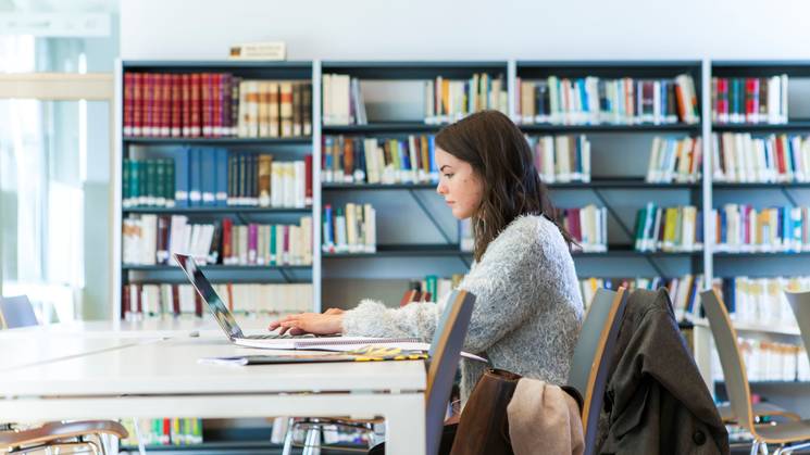 A woman is working on a laptop in a modern library setting.