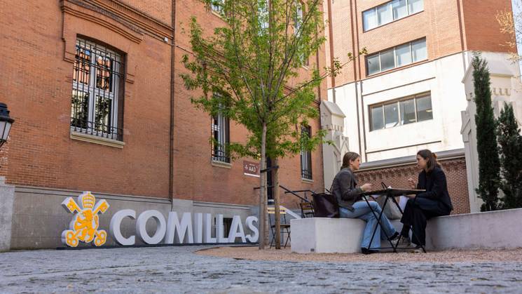 Dos personas conversan en un ambiente al aire libre frente a un edificio de ladrillo con un cartel que dice 'COMILLAS'.