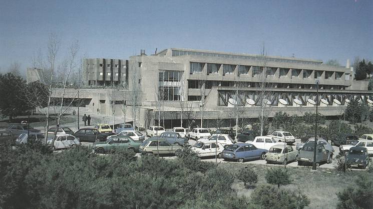 A vintage photo of a parking lot filled with cars in front of a multi-story building, likely a school or office building, under a clear sky.