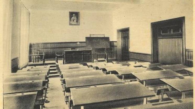 An old classroom featuring rows of wooden desks and a blackboard at the front, under a portrait.