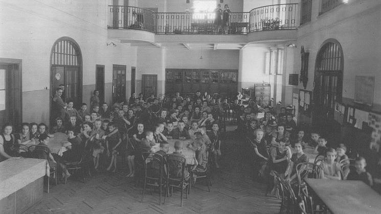 Una fotografía en blanco y negro de un gran grupo de niños en un salón con mesas y sillas.