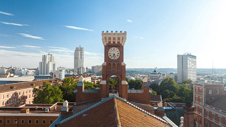 Reloj en torre de ladrillo en el campus universitario de Comillas con edificios y ciudad de fondo bajo cielo despejado