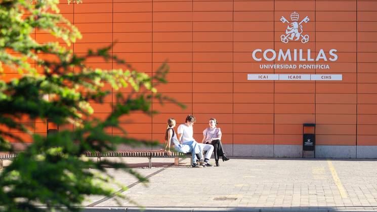 Tres estudiantes conversan sentados en un banco frente a la Universidad Pontificia Comillas.