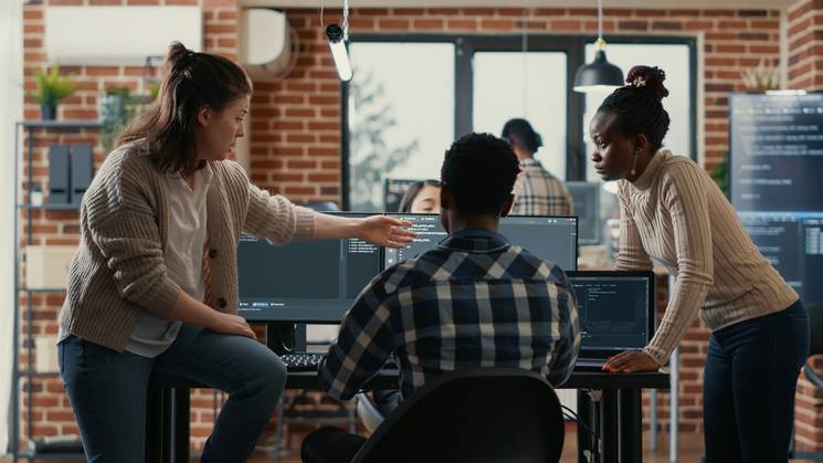 A diverse group of colleagues discussing work in a modern office setting with multiple computer screens.