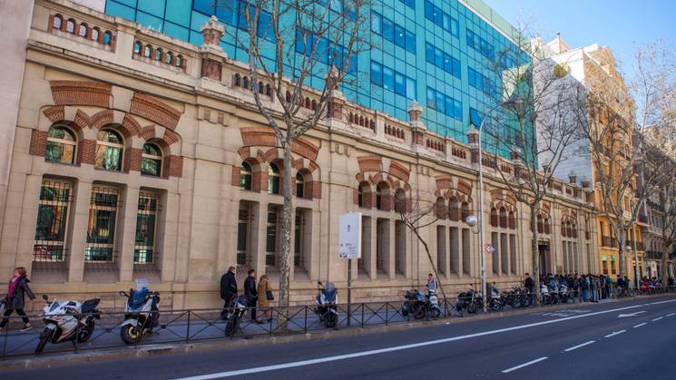 La imagen muestra un edificio histórico a la izquierda y una moderna estructura de vidrio a la derecha, con personas caminando y motocicletas estacionadas en la acera.