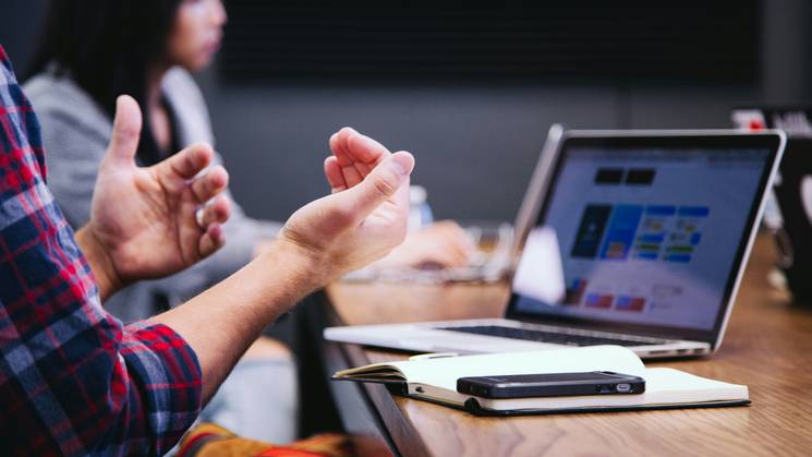 A person in a plaid shirt gesturing with hands during a discussion in a modern office setting.