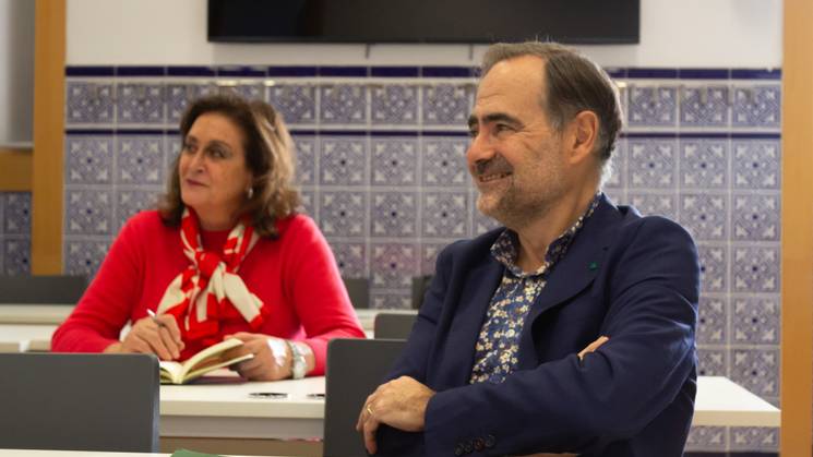 A man and a woman sitting at a table in a classroom, both looking towards the left with attentive expressions.