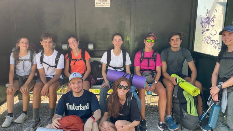 Un grupo de jóvenes con mochilas y esterillas esperan en una parada de autobús.
