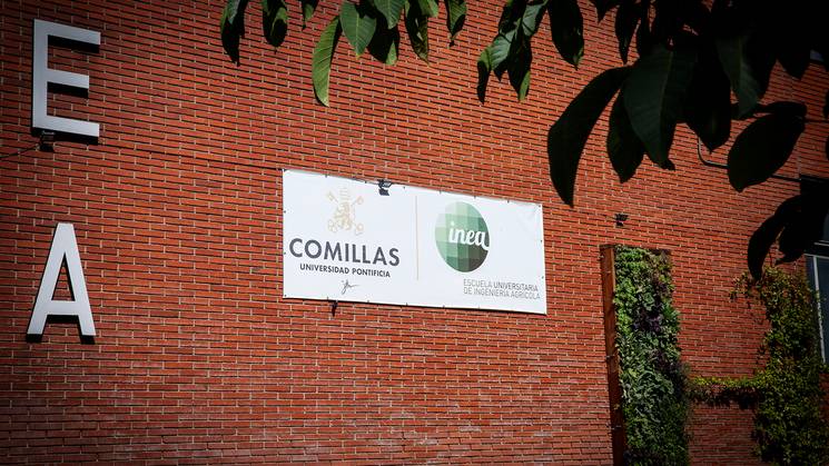 La imagen muestra un cartel de la Universidad Pontificia Comillas con el logotipo de INEA en una pared de ladrillo.