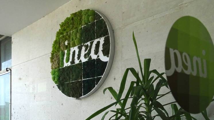 A circular green living wall with the word 'Inea' integrated into the design, displayed on a concrete wall next to a glass panel with reflections.