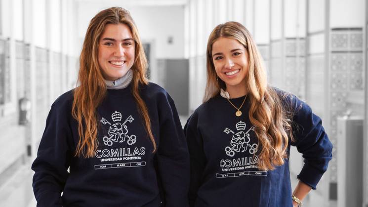 Dos estudiantes sonrientes posan en un pasillo de la universidad vistiendo sudaderas con el logo de Comillas.