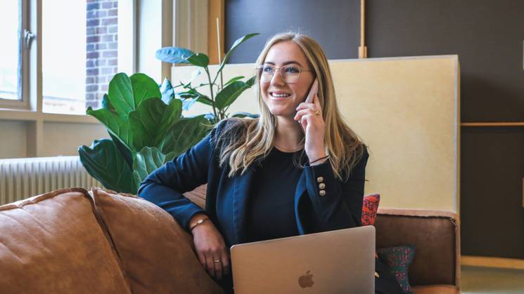 A woman in business attire is sitting on a couch, using a laptop and talking on the phone in a cozy office environment.