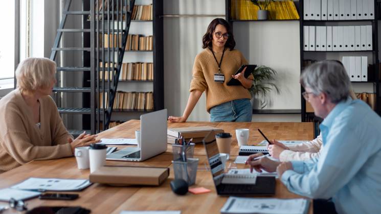 A young woman stands presenting in a boardroom meeting to two older colleagues who are taking notes.