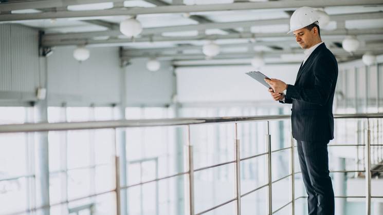 Un hombre vestido de traje y casco de seguridad revisa documentos en un entorno industrial.