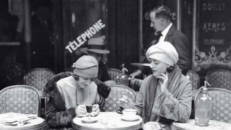 Dos mujeres sentadas en una terraza de café, vestidas a la moda de los años 20, disfrutando de una taza de café.