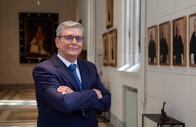 A man in a blue suit and glasses stands with his arms crossed in a corridor lined with portrait paintings.