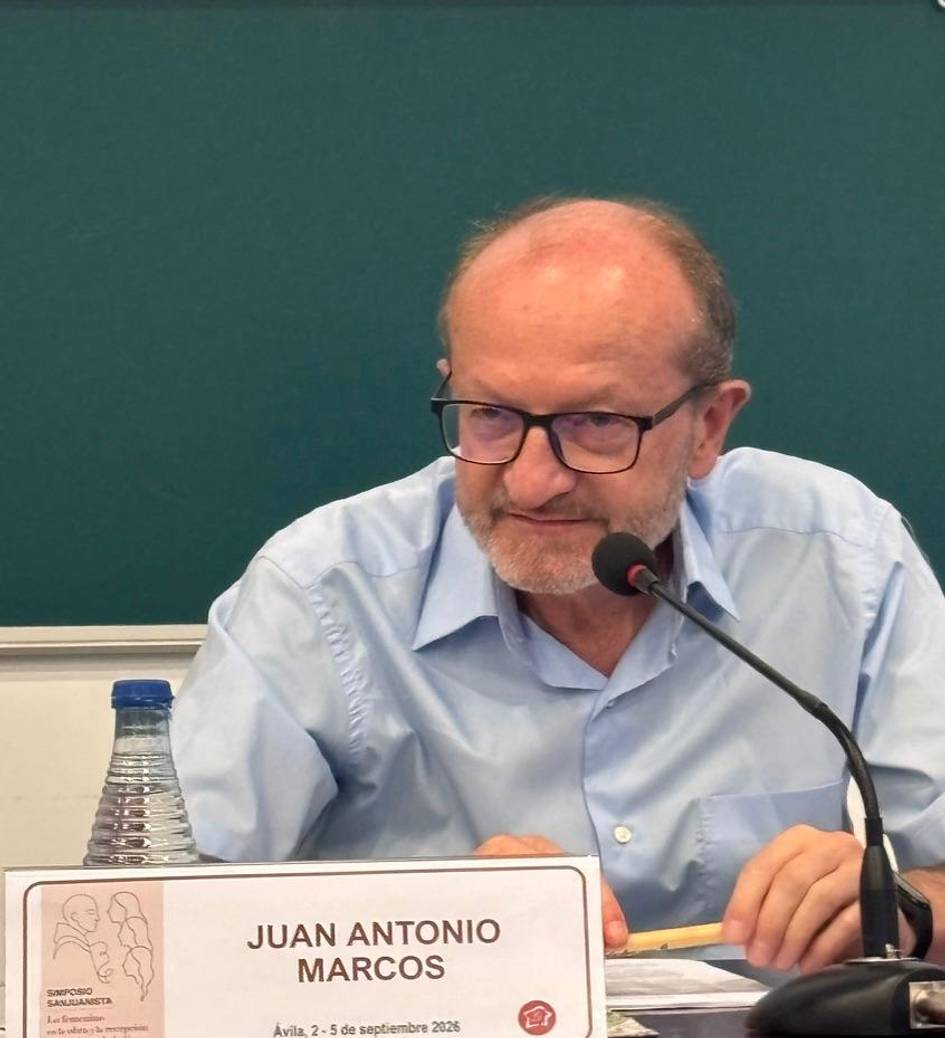 Un hombre con gafas sentado frente a un micrófono en un evento con un fondo verde.