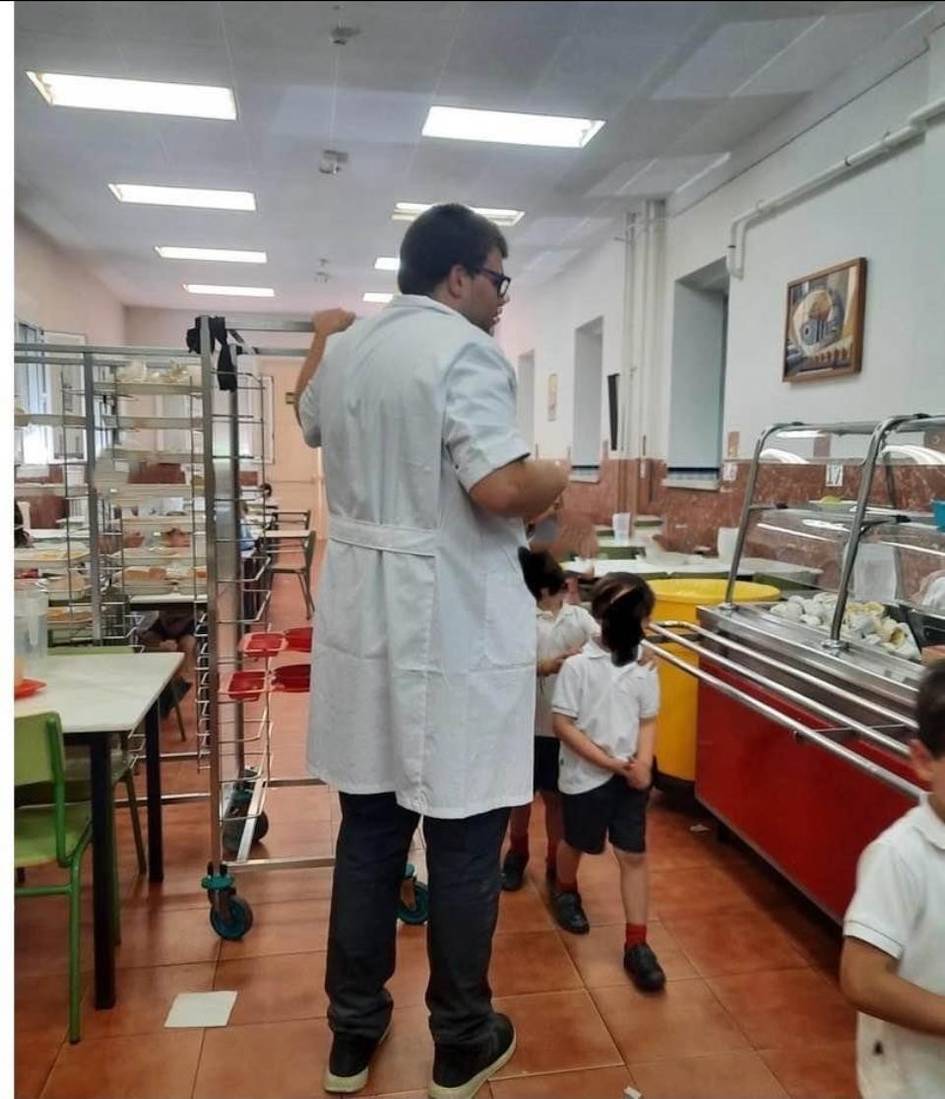 Un adulto de pie en un comedor escolar observando a unos niños pequeños en fila.