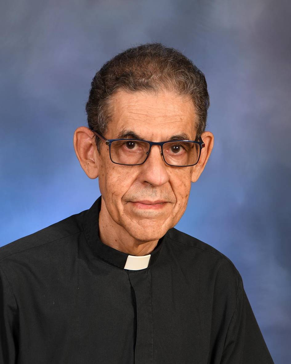 Portrait of an elderly priest wearing glasses and a clerical collar against a blue background.