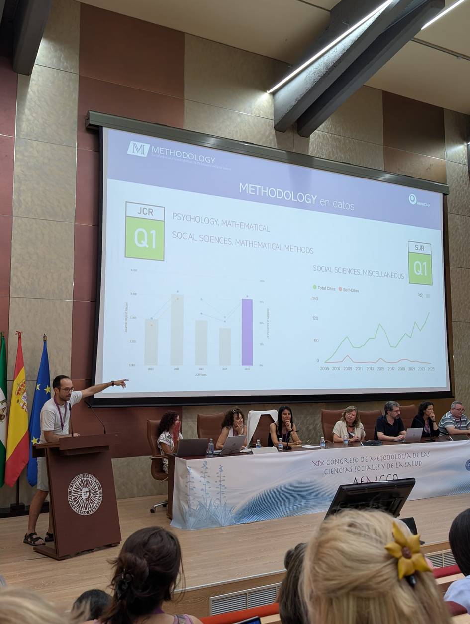 Un presentador está explicando gráficos sobre metodología en un congreso académico mientras un panel de ponentes lo observa.