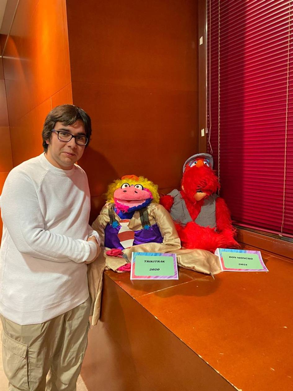 Un hombre posa sonriendo junto a dos marionetas coloridas, una morada y otra roja, sentadas en una mesa con carteles delante.