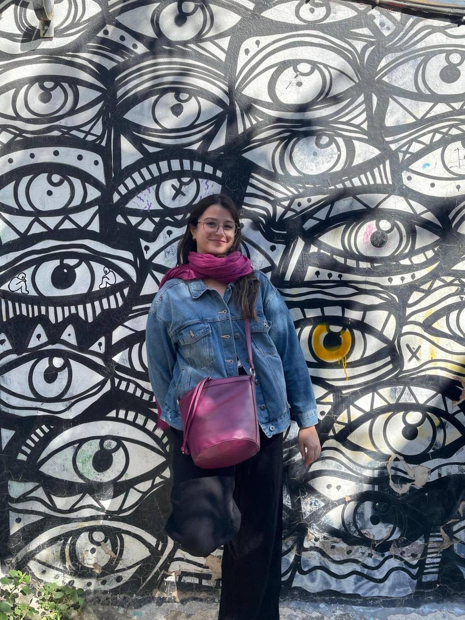 Una mujer posa frente a un mural de grandes ojos en una pared.