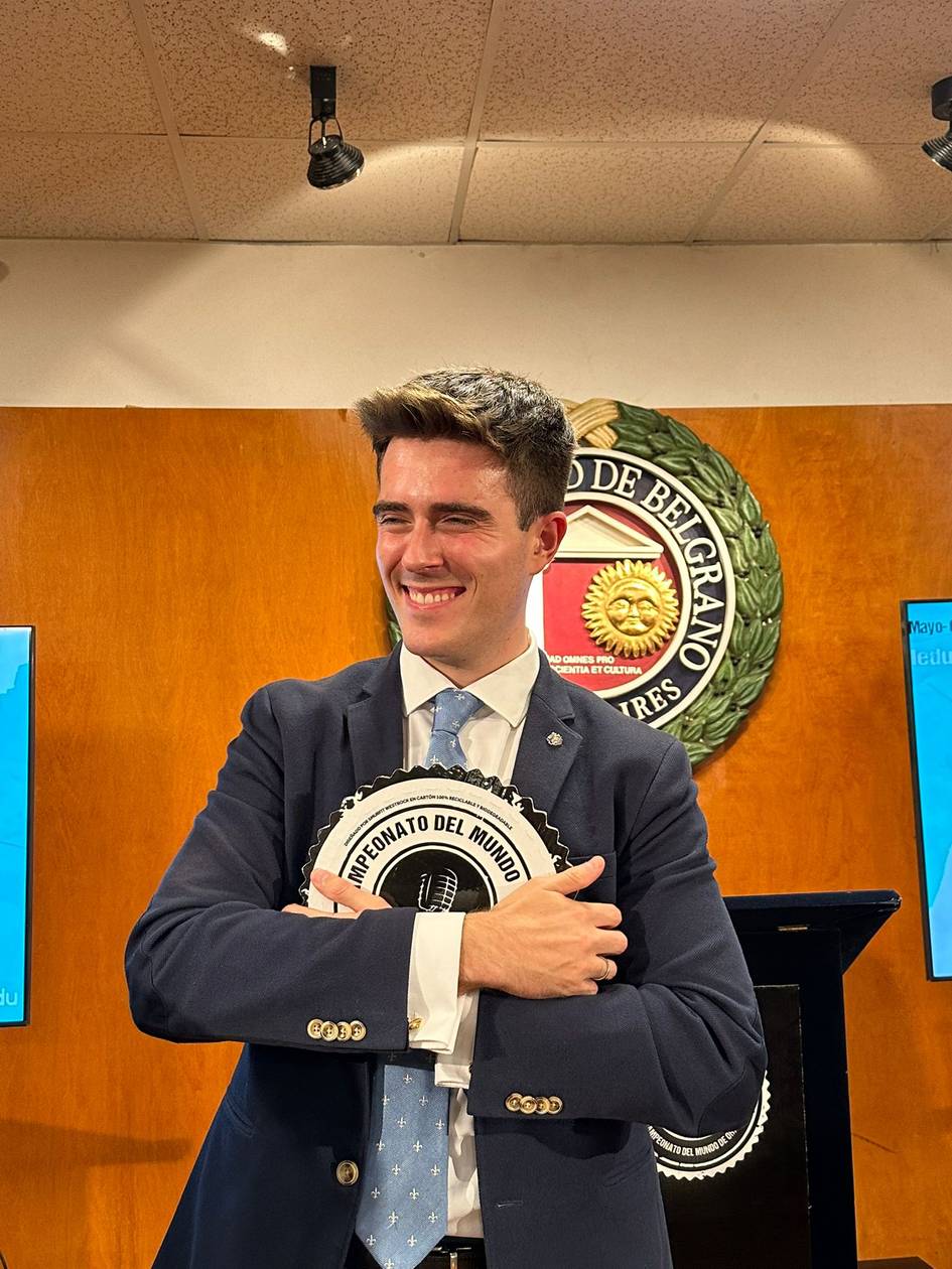 A smiling young man in a suit hugs a decorative plaque in a room with a wooden panel and a logo of 'Bacardi' behind him.