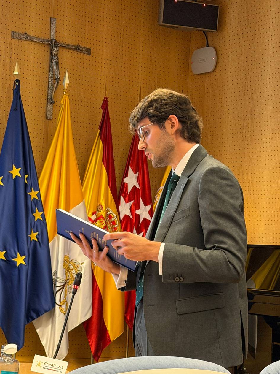 Un hombre en traje sostiene un documento mientras habla en una sala con banderas y un crucifijo de fondo.