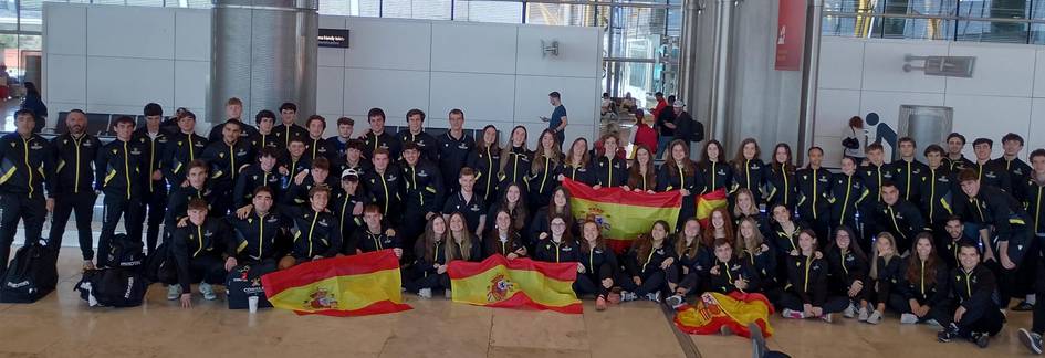 Grupo de jóvenes atletas con uniformes negros posando con banderas de España en un aeropuerto.