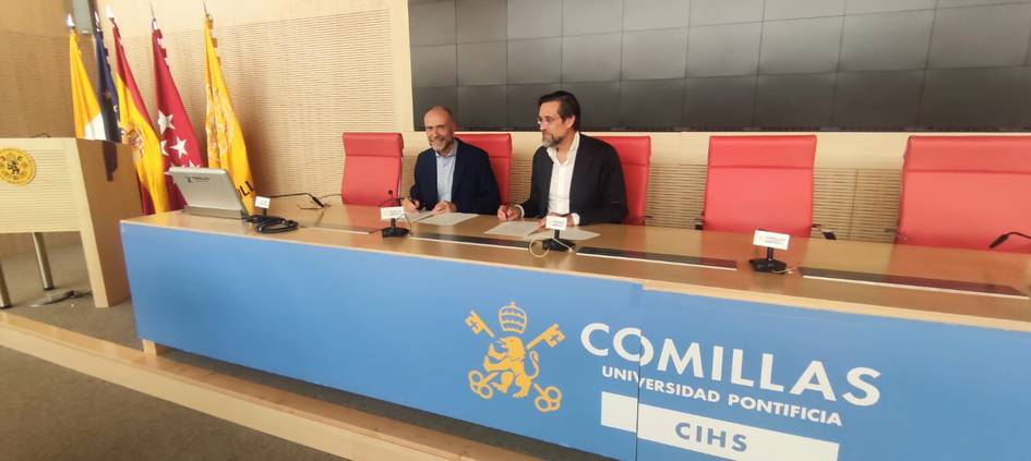 Two men sitting at a table during a conference at Comillas Pontifical University, with flags in the background.