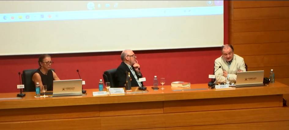 Three people sitting at a panel table in a conference room, engaged in discussion with laptops and microphones in front of them.