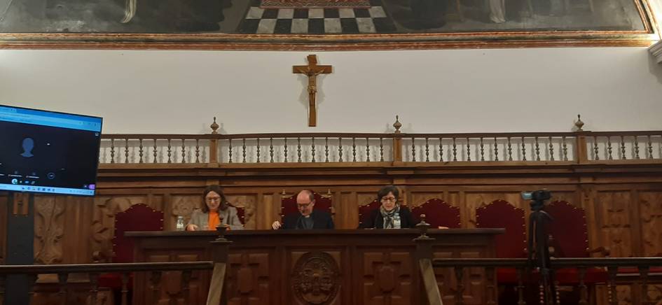 Three people sitting behind a long table in a panel setup with a crucifix on the wall above them in a room with ornate wooden decor.