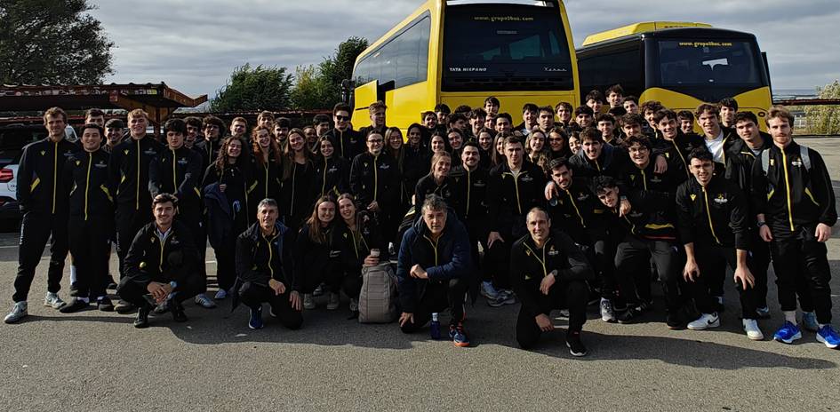Un grupo grande de jóvenes en uniforme deportivo se agrupan frente a dos autobuses amarillos.