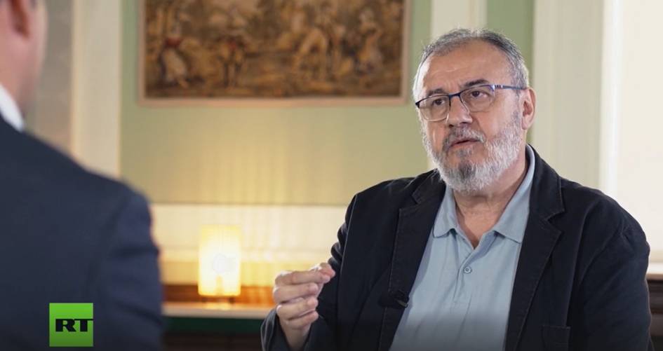 A man with glasses sitting and talking during an interview in a room with a painting on the wall.