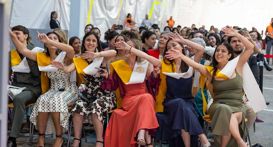 Un grupo de mujeres celebra con entusiasmo en un evento, vistiendo vestidos elegantes y bandas de graduación.