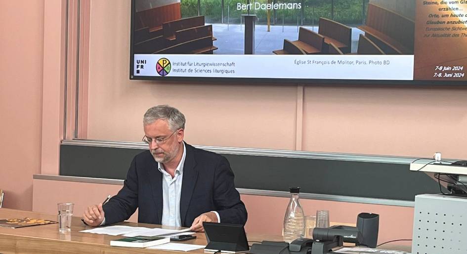 A man sitting at a desk with papers, speaking in a meeting room with a presentation in the background.