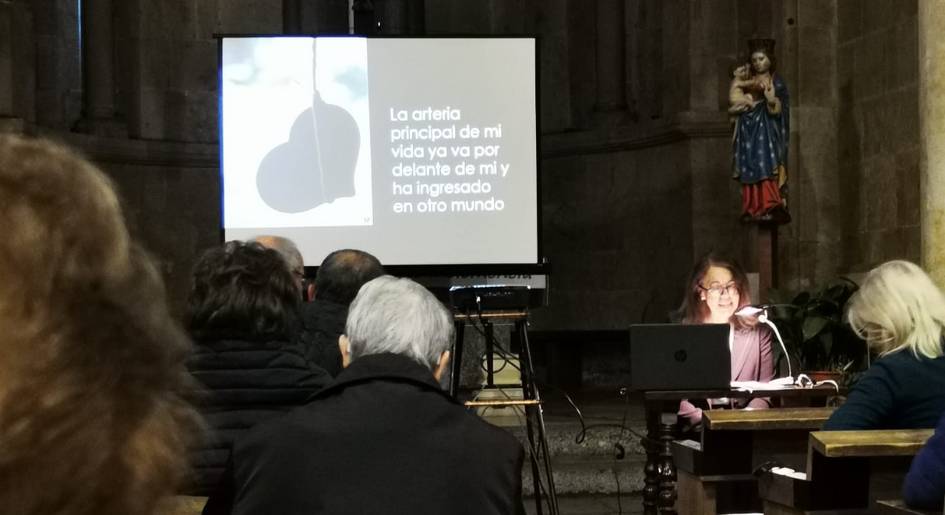 A woman presents a slide with text in Spanish during an indoor event, while attendees watch.