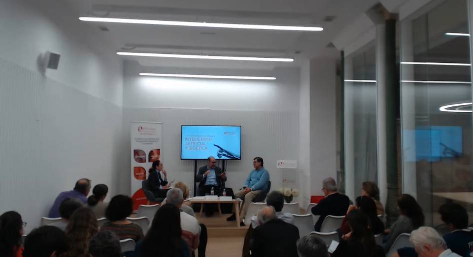 A panel discussion at an indoor event with three speakers sitting in front of an audience with a projector screen in the background.